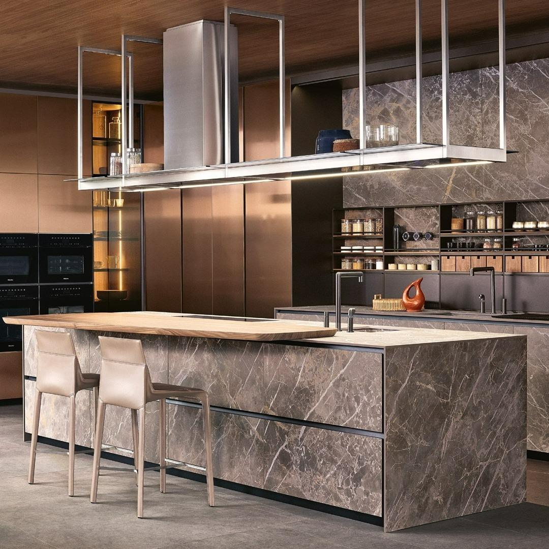 تصميم مطبخ حديث أنيق يتميز بجزيرة رخامية كبيرة مع طاولة إفطار خشبية مدمجة، وكراسي بار جلدية، وخزائن خلفية ذات ألوان برونزية دافئة.
